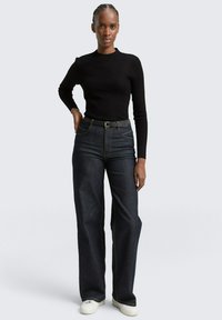 Schwarzes, geripptes Langarmoberteil kombiniert mit dunkelblauen, weiten Jeans und einem schwarzen Gürtel. Das Outfit beinhaltet weiße Sneakers und sichtbare Nähte.