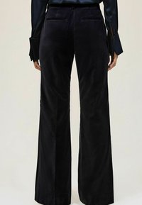 Persoon met zwarte fluwelen wijde broek en een donker shirt met lange mouwen, gezien vanaf de achterkant tegen een lichte achtergrond.