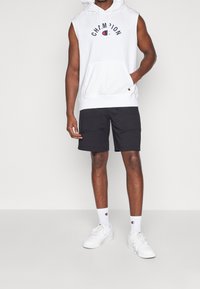 Uomo che indossa un hoodie senza maniche bianco Champion, pantaloni corti neri, calzini bianchi Champion e sneakers bianche, in piedi contro uno sfondo neutro.