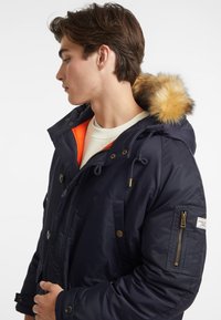 Chaqueta con capucha azul marino con detalles en naranja, borde de piel en la capucha, botones y cremalleras, dos bolsillos frontales y un bolsillo lateral en la manga con etiqueta.
