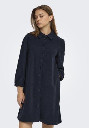 Femme portant une robe boutonnée bleu foncé à manches longues avec col, regardant vers le bas et sur le côté, sur un fond uni.