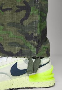 Vihreän sävyiset maastokuvioiset housut yhdistettyinä valkoisiin ja neonkeltaisiin tennareihin, joissa on tummansiniset korostukset ja kuvioidut pohjat.