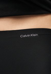 Κοντινό στο μέση ενός ατόμου που φοράει μαύρο εσώρουχο Calvin Klein με εμφανές λογότυπο της μάρκας στη ζώνη, πάνω σε γυμνό δέρμα.
