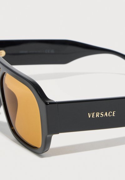 Μαύρα ορθογώνια γυαλιά ηλίου με κεχριμπαρένια φακούς και το λογότυπο της Versace σε χρυσό πάνω στον χοντρό βραχίονα του σκελετού, εμφανιζόμενα από πλάγια γωνία.