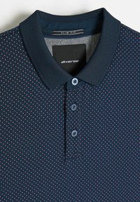 Granatowa, dopasowana koszulka polo z teksturowanym kołnierzem, ozdobiona wzorem małych różowych kropek. Trzy guziki z dopasowanymi guzikami.