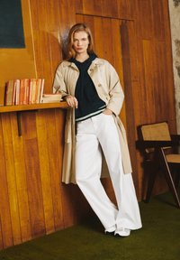 Un abrigo trench beige sobre una sudadera azul marino, pantalones anchos blancos y zapatillas. Fondo de panel de madera con libros en una estantería. Alfombra verde.
