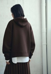 Sudadera marrón de gran tamaño con capucha, que presenta una textura suave y hombros caídos, superpuesta a una blusa blanca y una falda marrón con pliegues.