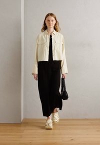 Cream cropped jacket over a black dress, paired with white slip-on clogs. The setting features a smooth gray wall and wooden floor.