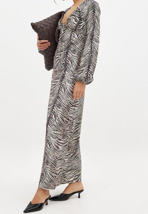 Mujer con un vestido largo de estampado de cebra con mangas abullonadas, sosteniendo un gran clutch tejido de color marrón oscuro y llevando tacones kitten negros con punta puntiaguda.