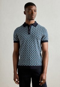 Granatowa koszulka polo w wzór z teksturowanym designem, wyposażona w czarny kołnierzyk i mankiety. Model zestawiony z ciemnymi spodniami, podkreślającymi dopasowanie koszulki.