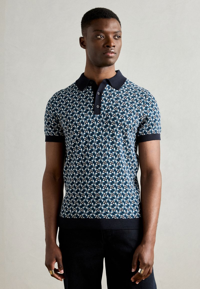 Granatowa koszulka polo w wzór z teksturowanym designem, wyposażona w czarny kołnierzyk i mankiety. Model zestawiony z ciemnymi spodniami, podkreślającymi dopasowanie koszulki.