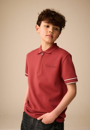 Jongen met krullend haar, gekleed in een rood poloshirt met korte mouwen en rits en een donkere broek, staand met één hand in zijn zak.