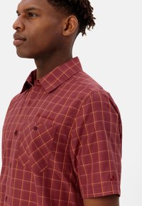 Kortotog shirt in bordeaux met een checkpatroon van oranje vierkanten, twee voorzakken en een rood logo op de borst. Zachte, gladde stoftextuur.