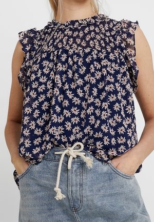 Kvinde iført navyblå ærmeløs bluse med hvidt margueritprint og lyseblå denimjeans bundet med et hvidt reb-bælte, hænderne i lommerne.