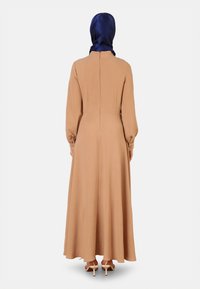 Long, flowy dress in beige with long sleeves and gathered cuffs. Back features a vertical seam. A blue satin hijab completes the look.
