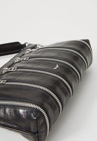 Sac en cuir noir avec des zippers en argent, présentant un design matelassé. Le sac dispose de plusieurs compartiments zippés et a une forme plate et rectangulaire.