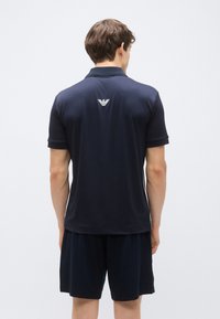 Temno modra polo majica z rebrastim ovratnikom in kratkimi rokavi, z belim logotipom na zadnjem delu ovratnika. Mehka, gladka tekstura tkanine.