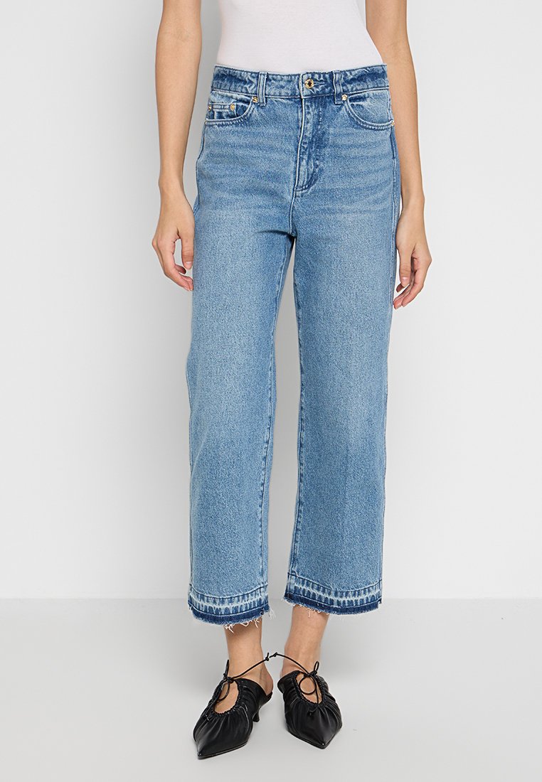 Jeans de pierna ancha en azul claro, fabricados en denim. Presentan una cintura alta, cinco bolsillos y un dobladillo deshilachado. Combinados con unos zapatos negros de tela, con recogido y sin cierre.