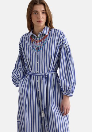Femme portant une robe chemise ceinturée à rayures verticales bleues et blanches, avec de longues manches bouffantes et des colliers en perles multicolores superposés.