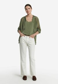 Camicia trasparente verde oliva con bottoni sopra un top coordinato, abbinata a jeans bianchi a gamba dritta e mocassini marroni.
