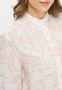 Blusa leggera e trasparente con un motivo floreale in morbido rosa e crema. Il design include un colletto alto e una chiusura con bottoni.