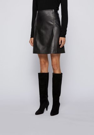 Kvinde iført en sort læder A-formet nederdel med lynlåslommer, kombineret med sorte ruskindsstøvler med hæl, der går til knæet, og en sort langærmet top.