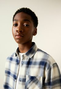 Blau-weiß kariertes Flanellhemd mit durchgehender Knopfleiste, Kragen und Tasche. Glatte Textur, getragen von einer jungen Person vor hellem Hintergrund.