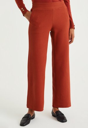 Woman wearing rust-colored textured wide-leg pants with hands in pockets and black loafers, standing on a light background.