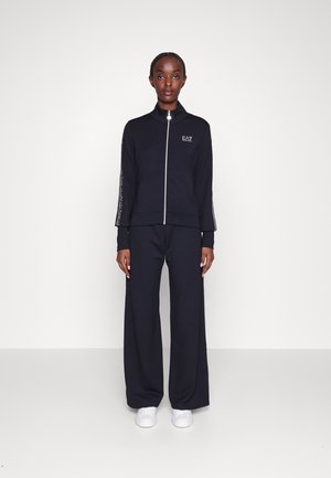 Giacca da ginnastica nera con zip e colletto alto, dotata di logo. Abbinata a pantaloni a gamba larga. Entrambi i capi hanno una finitura liscia. Sneakers bianche completano il look.