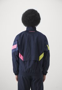 Marineblå træningsjakke med pink, blå og gule accenter, der har en høj krave og elastiske ærmer, letvægts, med en løs pasform.