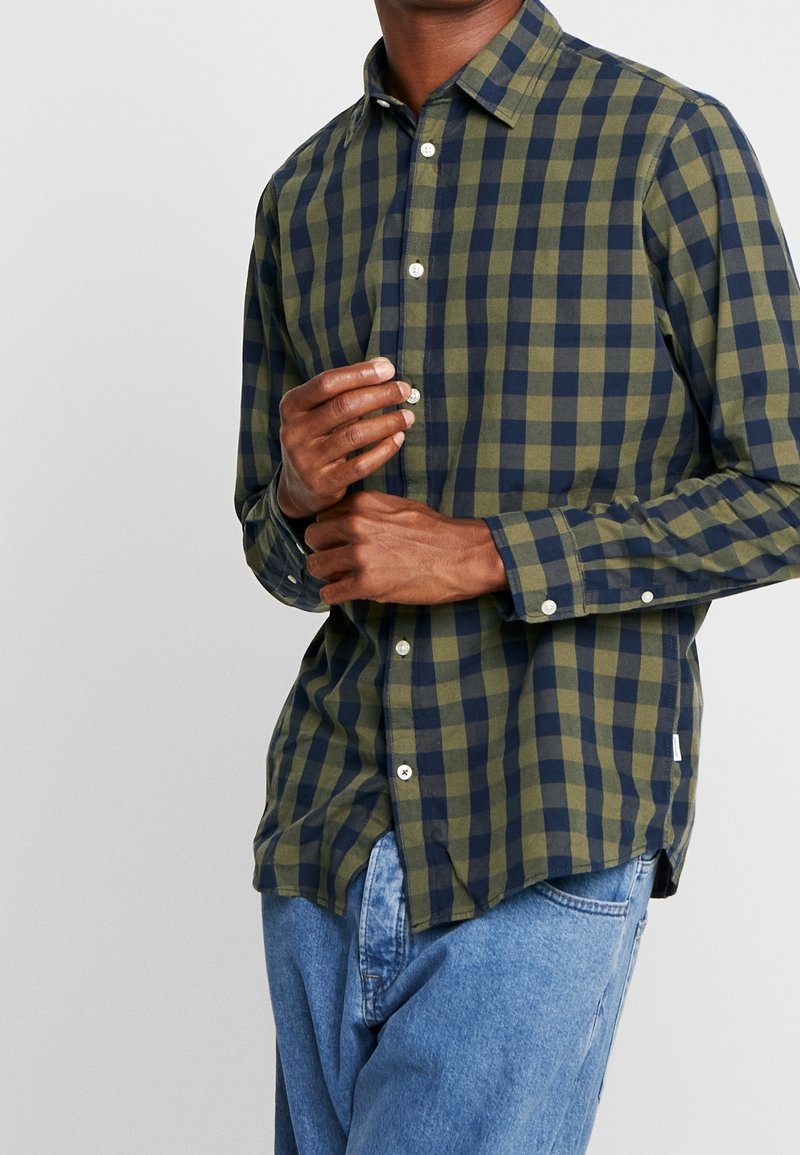 Personne boutonnant une chemise à manches longues à carreaux verts et bleu marine, portant un jean bleu clair, sur un fond gris clair uni.