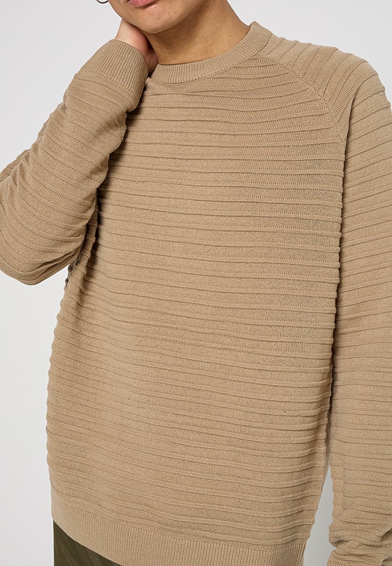 Suéter de punto beige con un patrón de canalé texturizado, escote de tripulación y mangas raglán. La tela suave ofrece un ajuste cómodo.