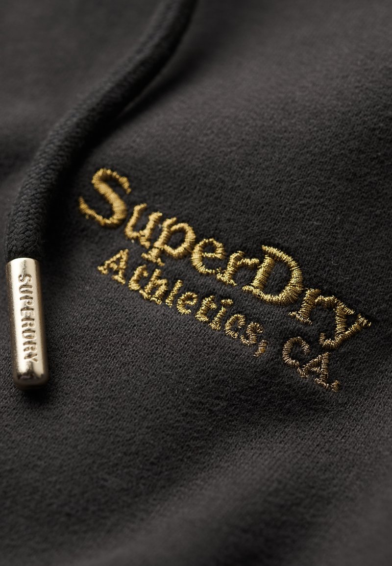 Schwarzer Kapuzenpullover mit goldenem, gesticktem "SuperDry Athletics, CA"-Logo. Verfügt über einen Kordelzug mit einemmetalenen Ende, das mit "SUPERDRY" graviert ist. Weicher Textil.