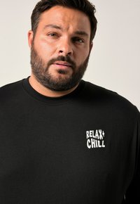 Černé tričko s dlouhým rukávem vyrobené z bavlny, s kulatým výstřihem a bílým textovým grafickým potiskem "RELAX + CHILL" na levé oblasti hrudníku.