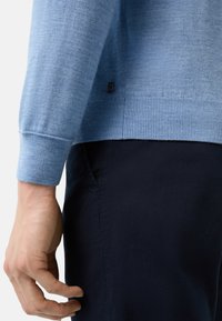 Hellblauer gestrickter Pullover mit geripptem Saum und Bündchen, versehen mit einem kleinen schwarzen Logo nahe dem Saum. Getragen mit dunkelblauen Hosen.
