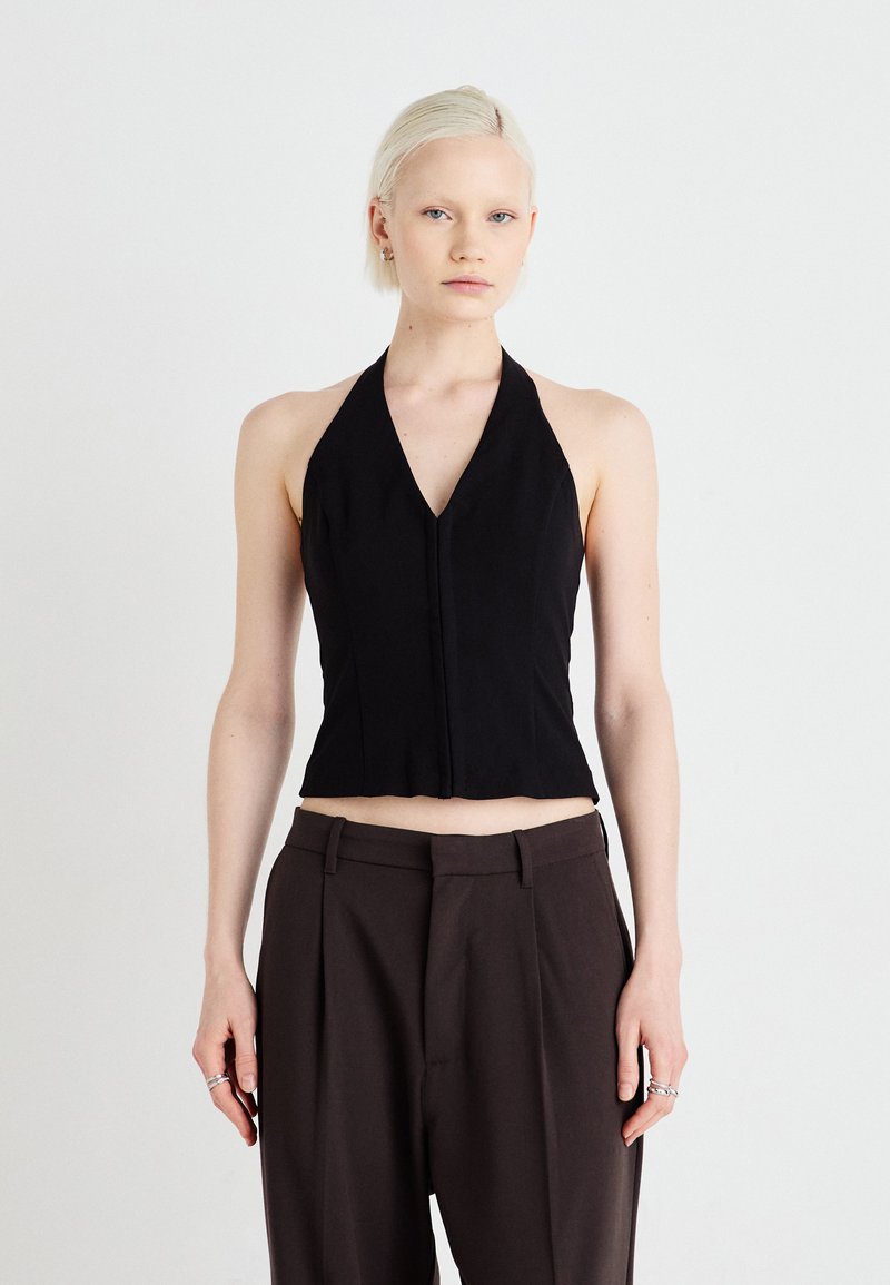 Black halter top with a deep V-neck and cropped design. Fabric appears smooth with minimal stitching. Worn with high-waisted brown trousers.