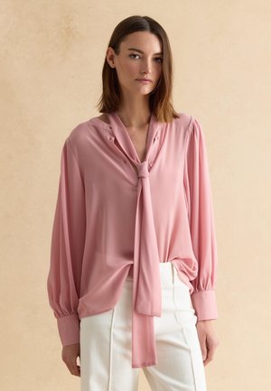 Donna con capelli castani lisci che indossa una camicetta rosa larga con fiocco al collo e pantaloni bianchi, in piedi davanti a uno sfondo beige.