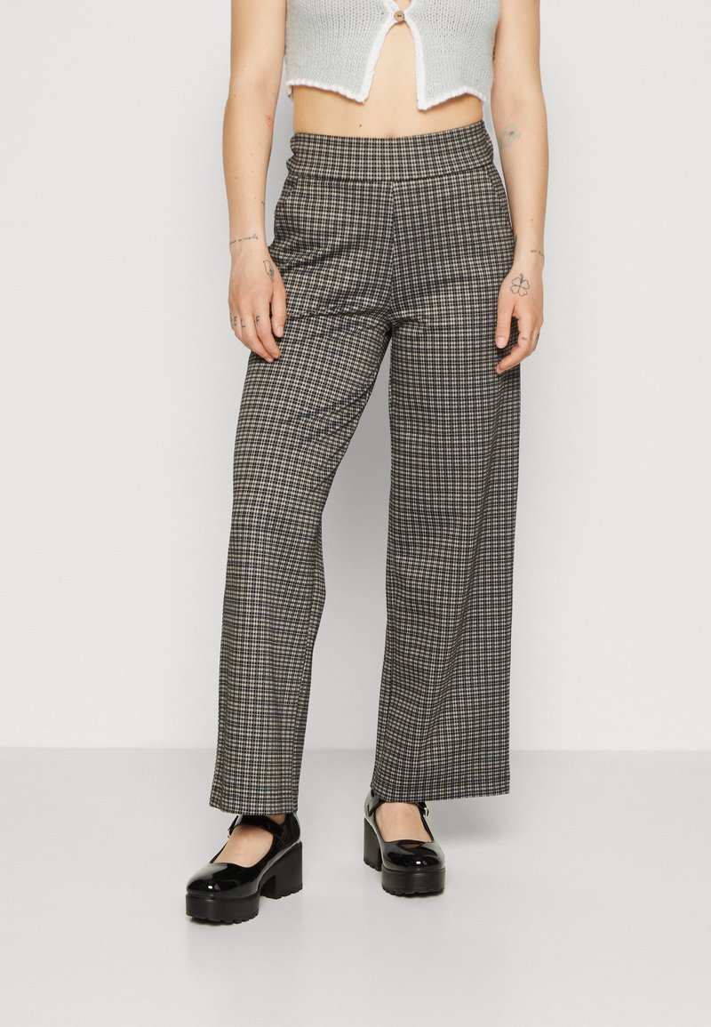 Personne portant un pantalon à carreaux taille haute et jambes larges, un haut en maille blanc court boutonné, et des chaussures Mary Jane noires à plateforme, debout sur un sol gris.
