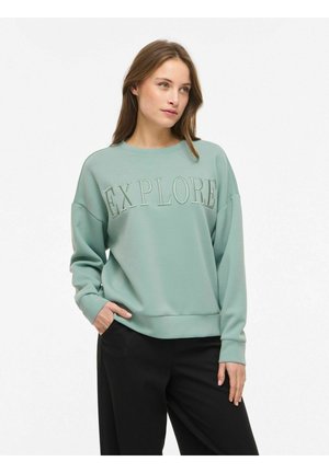 Jonge vrouw die een lichtgroene trui draagt met de tekst "EXPLORE", met één hand in de zak van een zwarte broek, kijkt iets opzij.