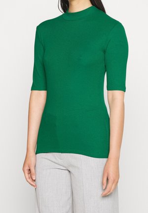 Femme portant un haut vert côtelé ajusté avec des manches aux coudes et un pantalon gris clair, debout devant un fond uni.
