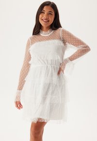 Vit tylklänning med prickigt mönster, volanger, långa ärmar och elastisk midja; lagerdesignen ger textur och visuell intressant.