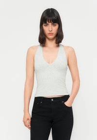 Kvinne med rett, mørkt hår som har på seg en lys grå halterneck-topp og svarte jeans, står med den ene hånden i lommen mot en enkel bakgrunn.