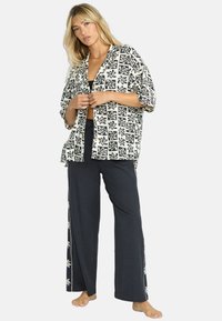 Camicia larga a righe bianche e nere con maniche oversize, indossata su una maglietta nera, abbinata a pantaloni blu navy con dettagli ricamati laterali.