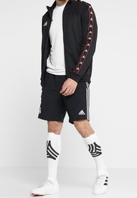 Chaqueta negra con cremallera y franjas laterales rojas, camiseta blanca, pantalones cortos negros, calcetines blancos con patrones negros y zapatillas de fútbol en un modelo.
