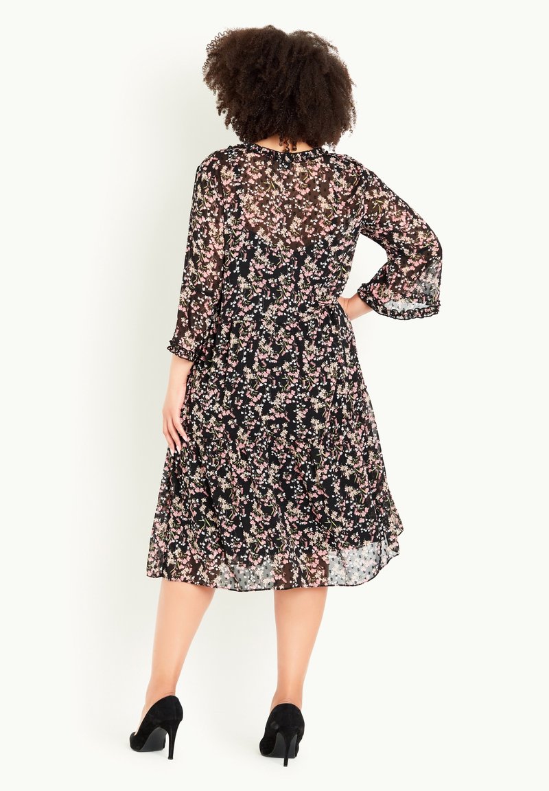 Femme aux cheveux bouclés portant une robe midi noire à fleurs avec des manches trois-quarts et des talons noirs, debout avec la main sur la hanche.