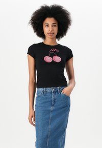 T-shirt nera aderente con design a ciliegia in strass rosa e testo "Guess" sopra. Il modello abbina una gonna in denim blu chiaro.
