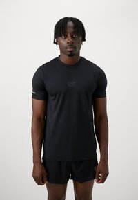 T-shirt de sport noir en matériau lisse et léger, à col rond, avec le logo EA7 sur la poitrine, porté avec un short assorti.