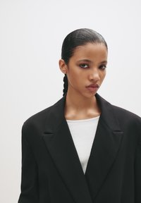 Femeie tânăr�ă cu părul strâns la spate, îmbrăcată cu un sacou negru peste un top alb, privind direct cu o expresie neutră pe un fundal simplu.