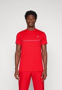 T-shirt rosso a maniche corte realizzato in tessuto morbido. Presenta una striscia orizzontale sul petto e un piccolo logo verde sul lato sinistro.