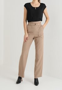 Top nero a maniche corte con dettaglio a chiave, abbinato a pantaloni beige a vita alta e gamba larga con cintura. Mule nere con tacco.